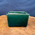 A small, 4.25-inch rectangular bonsai accent pot from the Seizan Kiln, featuring a deep emerald green glossy glaze. The ceramic pot has a deep, rectangular shape with subtle corner curves and sits on four small, low-profile feet. Its rich green surface has a slightly mottled, high-shine finish that reflects light. The pot is positioned on a polished, natural wood slab with a prominent, warm-toned grain. The background is a simple, dark blue textured surface, providing a stark contrast that highlights the pot's vibrant color and form. The unglazed interior of the pot is visible from the top, showing the natural clay body.