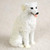 A small, detailed white Kuvasz dog figurine designed as a bonsai tree accessory or miniature garden ornament. The figurine depicts the Kuvasz in a sitting position with its head turned slightly to the side, featuring a cheerful expression with a pink tongue lolling out, dark expressive eyes, and a small black nose. The sculpture realistically captures the breed's characteristic thick, wavy white coat through intricate, textured grooves. This miniature white dog figurine is perfect for adding a lifelike touch to bonsai displays, fairy gardens, or indoor plant arrangements. The figurine is set against a soft, neutral, light-colored background that emphasizes its crisp white color and detailed craftsmanship.