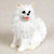 A detailed miniature figurine of a white American Eskimo dog, designed as a decorative accent for a bonsai tree arrangement. The small figurine is sculpted with a deeply textured coat to mimic thick, fluffy white fur. It is depicted in a sitting position with its head turned slightly to the left, featuring small black eyes, a black nose, and an open mouth with a tiny pink tongue showing. Its signature pointed ears have a subtle beige tint on the inner edges, and its characteristic bushy tail is curled against its side. The figurine stands out against a light, neutral-toned background, highlighting its bright white color and intricate details.