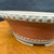 A detailed close-up, side-angle shot of an 8-inch Bigei Tokoname round bonsai pot. The pot is crafted from unglazed reddish-brown clay and features two distinct decorative elements: a thick beaded rim at the top and a matching, smaller-scale beaded foot band just above the base. 

A fine white patina or mineral dust settles in the crevices of the rounded beads, highlighting the intricate texture and giving the pot a classic, aged appearance. The pot has a tapered profile that narrows toward the bottom, resting on small integrated feet. It sits on a polished, warm-toned wooden surface against a soft, dark blue background that emphasizes the craftsmanship of the Japanese Tokoname pottery.