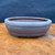 An authentic 6.25-inch Bigei Tokoname oval bonsai pot with a rounded band, crafted from high-quality, unglazed Japanese clay. The pot features a matte, dark charcoal grey to deep purple-brown finish, characteristic of traditional Tokoname pottery. Its elegant, shallow oval design is highlighted by a prominent raised horizontal band encircling the upper rim, with a subtle indentation near the base. The pot sits on small, integrated feet designed for stability and drainage, resting on a polished light brown wood-grain surface. The backdrop is a soft, dark blue, which emphasizes the fine texture and artisanal silhouette of this handmade bonsai container.