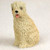 A small, hand-painted figurine of a Soft Coated Wheaten Terrier, designed as a decorative accessory for bonsai trees or miniature gardens. The figurine is captured in a sitting position, looking forward with a slight tilt of its head. Its coat is a pale cream or wheaten color, meticulously textured with wavy lines to mimic the breed's characteristic soft, curly fur. The dog's face features a prominent black nose, dark eyes peeking through its fringe, and a happy expression with its mouth open and a small pink tongue showing. The miniature sits against a plain, off-white background, highlighting the fine details of its craftmanship, from the tufts of hair on its head to its compact, seated form.