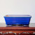 A rectangular ceramic bonsai pot sits centered on a dark wooden display stand. The pot is finished in a rich, vibrant cobalt blue glaze with a glossy sheen. A delicate network of fine crackle lines, or crazing, is visible throughout the blue finish, adding subtle texture. The pot features a flared, rounded top rim and sides that taper slightly toward the base. The interior of the pot is a contrasting pale cream color. It is elevated by small, integrated feet at each corner. The pot rests on a polished, dark mahogany-colored wooden table with ornate, scroll-like carvings along the front edge, some of which have reflective silver-colored accents. The background is a plain, neutral off-white wall, making the deep blue of the ceramic vessel stand out prominently.