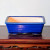 A front-view studio photograph showcases a vibrant cobalt blue ceramic bonsai pot resting on an ornate wooden surface. The pot is rectangular with gently rounded corners and sides that taper slightly toward the base. Its exterior is finished with a glossy, deep blue glaze that exhibits a subtle, fine-lined crackle or crazing pattern across its surface. The wide, prominent rim shares this same lustrous blue finish, while the pot's interior remains unglazed, revealing a clean, off-white or light beige ceramic texture.

Small, integrated feet at the bottom corners slightly elevate the pot, creating a narrow gap between its base and the furniture below. The pot is centered on a polished, dark reddish-brown wooden table or display stand, which features a decorative apron with intricate, hand-carved openwork designs along its bottom edge. The entire scene is set against a plain, neutral off-white wall, which provides a minimalist backdrop that highlights the rich color and craftsmanship of both the ceramic pot and its wooden pedestal.