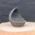 A handcrafted, dark slate-gray concrete planter with a unique, organic shape is positioned on a light-colored wood slab against a dark, textured background.

The planter resembles a broken eggshell or a hollowed-out geode. It features an asymmetrical design where one side rises into a tall, curved peak, while the opposite edge curves down into a lower rim. This creates a deep, protected interior space.

The exterior surface is rugged and craggy, with a coarse texture filled with pits and ridges that mimic the appearance of natural weathered rock or volcanic stone. In contrast, the interior is smoother but retains a fine, porous quality. The lighting highlights the planter's varied textures, casting soft shadows and bringing out light gray highlights along its irregular edges.

The planter sits in the center of a circular, light-toned wooden base with a natural grain. The background is a solid, dark charcoal-colored fabric, which provides a stark contrast that makes the sculptural form of the object stand out.