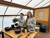 Two smiling beginner bonsai workshop students standing behind a classroom table with Jade bonsai trees, ceramic pots, bonsai soil, wire, tools, and planting materials. One student is holding a tree with visible roots while the other stands beside a partially styled Jade bonsai, showing the hands-on nature of the workshop.