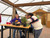 Bonsai workshop scene inside a greenhouse classroom showing a student leaning over a Jade bonsai tree while receiving hands-on guidance at a worktable. The table includes nursery pots, soil, tools, and workshop materials, with other students seated nearby in a bright greenhouse setting.