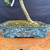 Close-up of the handmade glazed Hackberry bonsai pot showing blue-green glaze, raised etched branch artwork, mossy soil, and the lower trunk emerging from the container.