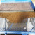 A detailed close-up shot of a blue industrial soil sifting machine in action, specifically processing hard premium Akadama bonsai soil. The top portion of the frame shows unrefined material on a coarse metal mesh. A horizontal black bar separates this upper stage from the lower section, where a large, centered mound of sifted, reddish-brown Akadama granules sits atop a fine blue mesh screen. The clay granules are clean, uniform in size, and noticeably free from the fine dust and small debris that often clogs soil. This professional sifting process is designed to ensure that only high-quality, dust-free granules remain, which is essential for providing the optimal aeration and drainage needed for healthy bonsai root systems. The clear contrast between the various mesh layers highlights the thorough cleaning and grading process.