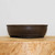 A high-quality, deep chocolate brown unglazed bonsai pot sits in a profile view on a thick, light-toned natural wood block. This classic oval bonsai pot features gently curved, convex sides and a distinct raised lip around the upper rim. Near the base, a subtle decorative horizontal band encircles the pot, just above its integrated small feet which provide essential elevation for drainage and airflow. The matte, earthy texture of the unglazed ceramic clay is clearly visible against the clean, neutral off-white background, highlighting its traditional aesthetic suitable for various bonsai tree styles.