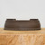 A professional studio photograph of a low-profile, 8.5-inch unglazed oval bonsai pot crafted from dark, chocolate-brown clay. The pot has a smooth, matte texture and an elegant, minimalist design. A distinct horizontal ridge or band encircles the upper third of the container, adding a subtle architectural detail. The top rim features a recessed inner edge with two symmetrical raised sections on the left and right sides, giving it a unique silhouette. The pot sits centered on a thick, light-toned wooden block with a prominent, natural grain, which provides a warm contrast to the dark ceramic. The entire composition is set against a clean, neutral white background, highlighting the earthy tones and refined shape of the bonsai container, making it ideal for a variety of coniferous or deciduous bonsai trees.