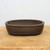 A professional-grade, unglazed oval bonsai pot crafted from dark brown ceramic with a subtle, matte earthy texture. The pot's design features a prominent rounded rim at the top and a decorative recessed band encircling the lower third of its body, adding a touch of elegance to its classic form. It rests on small, sturdy feet that provide slight elevation from the surface, essential for proper drainage. The pot is centered on a natural, light-colored wooden stump or table, set against a clean, minimalist off-white background. Soft, even lighting highlights the fine details and quality of the unglazed clay material, making it an ideal choice for various bonsai styles.