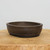 A professional-grade oval unglazed ceramic bonsai pot in a deep, earthy dark brown. This classic bonsai container is shown centered on a rustic, light-grained natural wood slice against a clean, off-white background. The pot features smooth, curved sides with a subtle horizontal line detailing near the rim and integrated drainage feet at the base to ensure optimal root health for a bonsai tree. The matte finish and simple design make it a versatile choice for a variety of bonsai species.