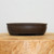 An eye-level, detailed photograph of a dark brown, unglazed oval bonsai pot resting on a light-toned wooden block. The pot features a low, elegant profile with a subtle, thin horizontal rim near the top and a similar recessed line along the base, creating a classic and balanced look. Small, discreet feet elevate the pot slightly, which is essential for proper drainage and aeration for a bonsai tree. The matte finish and fine-grained texture of the unglazed clay provide a natural, earthy aesthetic that complements the organic forms of a bonsai. Set against a clean, neutral off-white background, the image highlights the professional quality and minimalist design of this essential bonsai accessory.