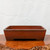 A front-facing view of a large, 15-inch rectangular unglazed Yixing bonsai pot. The pot is made from a rich, deep reddish-brown clay with a matte, finely textured finish and subtle light speckling. Its design is minimalist and sturdy, featuring clean lines and sides that slope slightly inward towards the base. Four integrated block feet are created by a wide, rectangular cutout along the bottom edge. The pot is shown resting on a polished, warm-toned wooden surface with a gnarled, natural-looking edge. The background is a simple, neutral white wall, highlighting the pot's earthy tones and geometric form. This classic Yixing clay pot is ideal for housing larger bonsai specimens.