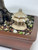 A close-up shot of a small, tan-colored Chinese pagoda figurine nestled on a bed of multicolored bonsai soil inside a dark brown ceramic pot. The miniature pagoda features two distinct tiers with white, tooth-like spikes along its roof edges and a rounded finial at the very top. It sits atop a variety of small rocks, including red lava rock and light-colored pumice, right next to the thick, gnarled trunk of a bonsai tree. The scene is set against a clean, plain white background, highlighting the intricate details of the bonsai decor.