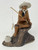 A detailed miniature Chinese clay figurine, commonly known as a "mud man," depicting an elderly man sitting serenely on a dark, craggy rock while fishing. The man is crafted from reddish-brown clay and features a long white beard, matching eyebrows, and a wide-brimmed white hat. He wears a simple brown robe and is shown in a relaxed pose with bare feet. In his hands, he holds a thin, light brown fishing rod with a white string line, at the end of which hangs a small, pale white fish. Beside his feet on the textured rock base sits a small, light gray woven-style basket or pot. This handcrafted bonsai accessory is presented against a plain white background, emphasizing its traditional design and fine details.