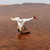 A detailed close-up shot of a miniature white crane figurine, a traditional Chinese mudman decoration designed for bonsai displays. The handcrafted ceramic bird is captured in a dynamic, graceful pose with its long neck extended forward and its wings partially outstretched as if landing or preparing for flight. 

The crane’s body is a textured matte white, featuring a striking bright red patch on the crown of its head and a long, pointed black beak. Subtle accents of dark brown and black are hand-painted onto the tips of the wings and tail feathers, adding a sense of realism and artisanal character. The bird is supported by two thin, dark brown wire legs that are anchored into a small, textured ceramic base resembling a weathered rock. 

The figurine is positioned on a polished, dark burl wood surface with rich, swirling grain patterns. The background is softly out of focus, transitioning from the warm tones of the wood to a neutral light grey, ensuring the delicate details of the crane remain the central focus.