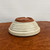 An upside-down view of a shallow, round, Japanese-style glazed bonsai pot showcasing its artisanal construction and functional design. The body of the pot features a cream-colored glaze with a speckled, crackled texture and subtle horizontal ridges that give it a handcrafted feel. The unglazed base, made of a warm reddish-brown clay, is prominently displayed at the top of the frame, revealing multiple pre-drilled drainage and wiring holes necessary for bonsai care. The circular foot rim is punctuated with three small notches for increased airflow and stability. The pot is set on a mottled, dark brown wooden surface against a plain, off-white background, highlighting its rustic elegance and specialized features for bonsai cultivation.
