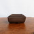 A set of three nested Udei brown unglazed bonsai pots in graduated sizes sits on a textured, warm-toned wooden surface. The rectangular ceramic pots feature a smooth, matte, dark chocolate-brown finish with slightly rounded corners and a subtle outward flare toward the top rim. Arranged from largest to smallest, the two inner pots are neatly tucked inside the front-most pot, which shows a small arched drainage cutout along its bottom edge. The pots are centered in the frame against a clean, minimalist off-white background, highlighting their earthy color and simple, elegant design.