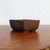 A medium-sized, rectangular Udei brown unglazed bonsai pot rests on a polished wooden surface with a visible grain. The ceramic pot features a minimalist design with a smooth, matte texture and a deep, chocolate-brown finish characteristic of unglazed clay. Its sides taper slightly inward toward the base, and the corners are gently rounded, giving it a soft yet structured appearance. A small semicircular drainage notch is visible at the bottom edge. The pot is photographed from a slightly low angle against a stark, clean white background, emphasizing its earthy tones and clean lines. This style of unglazed bonsai container is ideal for traditional bonsai presentations, offering a natural and understated aesthetic.