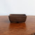 A rectangular Udei brown unglazed bonsai pot is centered on a polished wooden table against a plain white background. The ceramic pot has a deep, matte brown color with smooth, slightly sloping sides and rounded corners. The wooden surface below has a rich, mottled grain and a warm, glossy finish, reflecting a soft light onto the base of the pot. This type of unglazed bonsai pot provides a classic, earthy aesthetic, making it an ideal choice for a variety of bonsai tree species.