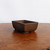 A rectangular, unglazed bonsai pot in a deep Udei brown color sits at a three-quarter angle on a textured, warm-toned wooden surface. The ceramic pot features a minimalist design with clean, straight lines and walls that taper slightly inward toward the base. Its matte finish highlights the natural, earthy texture of the clay. Small, arched drainage cut-outs are visible at the bottom edge, which also serve as subtle feet for the container. The pot is showcased against a plain, bright white background, emphasizing its traditional shape and rich brown hue, ideal for classic bonsai tree styling.