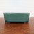 A handcrafted Japanese Tokoname bonsai pot with a classic rectangular shape and elegantly notched corners. The exterior is coated in a vibrant, glossy teal or seafoam green glaze that features a subtle, fine-speckled texture. The glaze darkens slightly near the top rim and bottom edge, creating a sense of depth and character. The bottom section of the pot is unglazed, revealing the dark, chocolate-brown color of the natural clay, and it rests on four small, sturdy feet designed for drainage. The pot is centered in the frame, sitting on a polished, warm-toned wooden surface against a minimalist, textured off-white wall background.