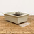 An elegant, cream-glazed rectangular Japanese Tokoname bonsai pot sits on a rustic, weathered wooden tabletop. The ceramic pot features a smooth, high-gloss cream finish that coats the exterior walls, stopping just above the unglazed dark brown clay at the base and feet. Its design includes a wide, flared rim and a decorative horizontal recessed line running across the middle of the container's sides. The pot rests on four sturdy corner feet, providing space for drainage and airflow beneath the vessel.

Inside the pot, a coil of dark brown bonsai training wire is visible. To the right of the pot on the wooden surface are two small, white square plastic drainage mesh screens, commonly used to prevent soil from escaping through the bottom holes. The entire scene is set against a clean, neutral off-white background, highlighting the minimalist aesthetic and artisanal craftsmanship of the traditional Japanese pottery.