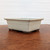 An elegant, side-angle photograph of a Tokoname cream-glazed rectangular bonsai pot, showcased on a polished, warm-toned wooden surface. The pot features a smooth, off-white glaze with subtle dark accents along the flared rim and edges, hinting at its handcrafted quality. A decorative horizontal line is recessed into the center of its tapered sides, providing a refined architectural detail. The bottom of the pot transitions into a dark, unglazed ceramic base with four sturdy, integrated feet, characteristic of traditional Japanese bonsai pottery. The minimalist background consists of a plain white wall with soft horizontal textures, focusing all attention on the craftsmanship and serene aesthetic of the ceramic vessel.