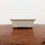 A high-quality Tokoname Japanese bonsai pot, featuring a rectangular design with a smooth cream-glazed finish. The pot has an elegant, outward-flaring rim and a subtle horizontal ridge around its body, highlighting its traditional minimalist craftsmanship. The base is left unglazed in its natural dark brown clay, showcasing sturdy cutout feet that provide stability and excellent drainage. It is displayed on a polished, reddish-brown wooden surface against a clean, textured white background, emphasizing the pot's refined aesthetic.