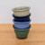 A vertical stack of four small, decorative glazed ceramic bonsai pots is arranged on a light-colored wooden tabletop against a clean white background. The collection showcases a variety of colors, featuring—from top to bottom—a cream-white, a dark navy blue, a soft periwinkle blue, and a muted sage green pot. 

Each pot is finished with a glossy glaze and distinctive design elements, including a vertically fluted body and a decorative scalloped rim. The bottom-most green pot rests on small, unglazed reddish-brown clay feet, highlighting the material and craftsmanship of these round bonsai containers. The image highlights the different color options available for this specific style of fluted ceramic pot.