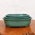 A set of three handcrafted Japanese Tokoname bonsai pots featuring a rich, glossy Oribe teal-green glaze. The pots are presented as a nesting set, graduated in size and stacked neatly inside one another, with the largest pot on the bottom and the smallest on top. 

Each container is rectangular in shape with soft, rounded corners and a slightly flared rim that is accented with a delicate, dark-toned border along the upper edge. The deep green glaze has a smooth, lustrous finish with subtle variations in color, characteristic of traditional Oribe-style Japanese pottery. 

The pots are resting on a highly textured, reddish-brown wooden burl slab that shows natural grain and knots, providing a rustic contrast to the elegant ceramic. The entire arrangement is set against a clean, minimalist off-white background that emphasizes the vibrant color and clean lines of the pottery. This set of Oribe glazed pots represents high-quality Japanese craftsmanship intended for displaying and cultivating bonsai trees.