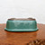 An upside-down view of a rectangular Oribe glazed Tokoname bonsai pot, showcasing its glossy teal or turquoise finish and precise craftsmanship. The ceramic pot features elegantly notched corners and a flared rim. The unglazed bottom of the pot reveals four sturdy corner feet and visible drainage holes, essential for bonsai health. The pot rests on a polished, natural-edge wood display stand with a rich brown grain, set against a clean, white background.