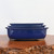 A stack of three graduated Japanese Tokoname bonsai pots sits centered on a rustic wood slab against a clean, minimalist white background. The pots feature a vibrant, glossy Namako glaze in a rich, deep cobalt blue that reflects light across its smooth surface. Each container is rectangular with softly rounded corners and a slight outer lip along the top rim. The largest pot on the bottom is the deepest, resting on small, unglazed ceramic feet that provide drainage clearance. Two shallower pots are nestled perfectly inside. The dark, polished wooden display stand has a rugged, organic "live edge" with natural burl textures, creating a striking contrast with the refined elegance of the blue glazed ceramics. This set exemplifies the high-quality craftsmanship and classic aesthetic of authentic Japanese bonsai containers.
