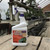 A white spray bottle of Bonide Liquid Copper Fungicide stands prominently in the foreground on a rustic wooden plank table. The bottle features a purple trigger and a label that indicates it is a ready-to-use solution for organic gardening, designed to control various plant diseases such as powdery mildew, black spot, downy mildew, and early blight. 

The background shows a bright, airy greenhouse or plant nursery. Several wooden tiered benches are lined with an assortment of bonsai trees in various ceramic pots, some with pink blossoms. The translucent greenhouse roof and structural metal supports are visible, along with black watering hoses coiled on the wooden walkway in the distance. This setting emphasizes the product's application for maintaining the health and beauty of delicate plants like bonsai.