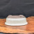 An upside-down view of a cream-colored oval glazed bonsai pot, revealing its unglazed tan underside. The pot features two large circular drainage holes and four integrated feet for proper aeration and drainage. It sits on a polished, natural-edge wooden display board against a textured black background, emphasizing the elegant contrast between the smooth cream glaze and the organic wood grain.