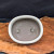 A top-down view of a cream oval glazed bonsai pot, resting on a polished live-edge wood slab with intricate grain patterns. The ceramic pot features a clean, off-white glazed rim that surrounds a matte, grayish-brown unglazed interior. Two prominent circular drainage holes are visible at the bottom, designed to ensure proper water flow and root health for a bonsai tree. The pot's minimalist aesthetic is showcased against a dark, solid background, highlighting its smooth contours and functional design for bonsai cultivation.