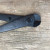 A detailed, close-up photograph from a top-down perspective showing the head and upper handles of a pair of black bonsai wire cutters. The tool is crafted from blackened carbon steel with a subtle matte finish and some visible oil marks or minor wear.

On the side of the lower handle, near the central pivot point, the name "YOSHIAKI" is clearly engraved in capitalized sans-serif letters. The pivot itself is a prominent circular rivet that joins the two arms. On the upper handle, just before the pivot, there is a small, raised cylindrical metal stopper designed to limit the tool's closing range.

The cutting head of the tool is rounded and robust, featuring two short, thick blades that meet at a precise, fine line. The blades are currently closed. The tool lies diagonally across the frame, resting on a weathered, light-brown wooden surface with a distinct horizontal grain pattern, punctuated by fine cracks, small pits, and a few tiny white flecks.