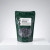 A front-facing studio product photograph of a dark green, resealable stand-up pouch containing Tinyroots Omakase Pellet Fertilizer for bonsai trees. 

The pouch sits on a clean, reflective white pedestal against a soft, light grey gradient background. The white label prominently features the brand name "Tinyroots" in the upper right and "OMAKASE Pellet Fertilizer" in bold black text. Below the name, the NPK ratio of 4/0/7 is displayed, alongside a black silhouette illustration of a classic bonsai tree on the left. 

A large, circular transparent window in the center of the bag reveals the contents: dark grey, textured, pill-shaped slow-release fertilizer pellets. The label includes detailed instructions for use, explaining how the pellets provide a continuous nutrient release over two months. This specialized Japanese-style fertilizer is designed for easy application across the soil surface of bonsai plants.