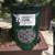 A front-facing, close-up shot shows a dark green, resealable pouch of Tinyroots Omakase Pellet fertilizer resting on the edge of a bonsai pot. The white label on the bag features the "OMAKASE" brand name in bold black letters, with an NPK ratio of 4/6/2 listed below. A stylized black silhouette of a bonsai tree is positioned to the left of the text.

Detailed directions on the label explain that the slow-release pellets provide continuous nutrients for up to two months when scattered across the soil surface and watered. A clear circular window at the bottom of the bag reveals the product's contents: large, oblong, dusty-gray organic pellets that resemble compressed earth. 

The fertilizer bag is propped up against a thick, gnarled trunk of a pine bonsai tree. The tree is planted in a shallow, reddish-brown ceramic pot filled with a gritty, multi-colored bonsai soil mix of pumice, lava rock, and akadama. In the soft-focus background, other bonsai pots and a wooden deck are visible, suggesting an outdoor garden or nursery setting.