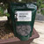 A dark green stand-up pouch of Tinyroots Omakase Japanese pellet fertilizer leaning against the textured trunk of a mature coniferous bonsai tree. The white label on the bag features a black bonsai silhouette and the product name "OMAKASE Pellet fertilizer" with an NPK ratio of 4/6/2. Through a clear circular window at the bottom of the bag, large, round, dark grey compressed organic pellets are visible. The pouch sits inside a rectangular brown ceramic bonsai pot filled with a professional, multi-colored gritty soil mix. The background shows a sunlit wooden deck and the soft-focus foliage of other bonsai trees.