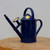 A side-profile view of a royal blue Haws indoor watering can resting on a thick, natural wood pedestal. The watering can features a classic, elegant design with a long, slender spout extending upwards for precise watering. A removable brass spray rose is conveniently parked on a storage peg near the base of the spout. The cylindrical body of the can is decorated with a raised diamond-grid pattern and has a small, circular gold brand medallion on its conical top. It is designed with both a tall, rounded top handle and a curved rear handle to provide balanced control while pouring. The background is a clean, solid light gray, which emphasizes the watering can's smooth, glossy finish. This 2-pint capacity tool is ideal for the delicate care required for bonsai trees and other indoor plants.