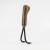 A two-tine garden hand cultivator stands upright against a clean, light gray studio background. The tool features a short, ergonomically shaped handle made of polished light brown wood with a dark, prominent grain pattern. A shiny silver metal ferrule, or collar, connects the base of the handle to the metal working end. Two sturdy black metal tines emerge from the collar, curving downwards and then sharply forward to end in pointed tips. The tool is balanced on these two tips, which are angled slightly towards the right. The lighting is soft and even, casting a faint shadow on the surface beneath the tines, highlighting the smooth texture of the wood and the matte finish of the metal.
