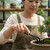 A close-up photograph shows a woman wearing an olive-green apron over a cream-colored long-sleeve shirt as she carefully tends to a bonsai tree. Her right hand is in sharp focus, holding the wooden handle of a two-pronged metal rake tool, which she uses to work the dark, pebbly soil at the base of the plant. The small bonsai tree, with its dense green needles and mossy base, is seated in a shallow, light-brown ceramic pot. The background is softly blurred, showing wooden shelves filled with various ceramic pots and greenery, creating the atmosphere of a serene greenhouse or garden studio.