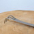 A detailed close-up shot showing the three-pronged rake end of a Tinyroots bonsai root rake and spatula tool. Made from sleek, matte-finished stainless steel, the tool’s three sharp tines are bent at a ninety-degree angle, designed to easily comb through soil and roots. The rake is positioned horizontally across a light-colored, natural wood surface with visible grain and texture. The clean, minimalist background is a soft, pale blue-gray, emphasizing the tool's professional quality and utilitarian design. This specialized bonsai tool is used for carefully untangling roots and clearing away old soil during the delicate process of repotting a bonsai tree.