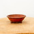 A small, unglazed Yixing bonsai pot made from reddish-brown clay sits centrally on a light-colored natural wood surface. The pot has a shallow, round bowl-like shape with a scalloped or lobed decorative rim. It is supported by three small, ornate feet with a subtle cloud-like design. The matte finish of the earthy clay is highlighted by soft, even lighting against a minimalist off-white background, emphasizing its simple and elegant craftsmanship.
