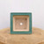 A top-down perspective of a square-shaped glazed ceramic bonsai pot, highlighting its functional design. The wide, flat rim is finished in a smooth, teal-green glaze with subtle variations in tone. The interior of the pot remains unglazed, showing the natural light-tan ceramic clay. 

The bottom of the pot features five pre-drilled holes essential for bonsai care: a large central drainage hole to prevent root rot and four smaller corner holes intended for securing the tree with bonsai wire. The pot is resting on a rustic, circular wooden display stand with a visible grain, all set against a clean, minimalist white background. This medium-sized container is designed for both aesthetic appeal and the specific health needs of a growing bonsai tree.