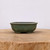 A detailed eye-level shot of a 4-inch glazed Yixing bonsai pot (No. 900) in a muted, mossy green color with fine dark speckles across its semi-matte surface. The pot features a rounded, oval-like shape with a gently inward-sloping body and a thick, smooth rim. It is elevated by small, integrated feet at the base. The pot is centered on a thick, rustic wooden block with a natural grain, which sits against a clean, minimalist off-white background. The soft lighting highlights the earthy tones and ceramic texture of the pot, emphasizing its simple yet elegant design.