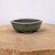 A 4-inch green glazed Yixing bonsai pot sits centered on a rustic, circular wooden slab. The small ceramic container features a soft-cornered rectangular shape with a prominent rounded rim and a muted, earth-toned green glaze that has subtle, dark speckles throughout. A slight vertical indentation is visible on the front side, adding a decorative touch to its form. The pot's dark, unglazed interior is visible, showcasing the natural texture of Yixing clay. It is elevated by small, integrated feet that provide a slight lift from the wooden surface. The wooden base shows a natural grain and light surface wear, set against a clean, neutral white background that highlights the pot's organic colors and handcrafted quality.